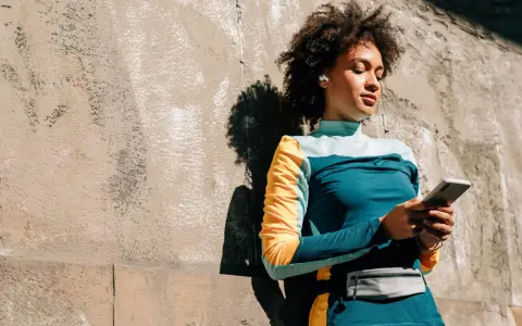 Frau mit Kopfhörern lehnt an Wand, hält handy beim joggen. Musik und Motivation für die Laufstrecke.