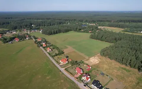 Widok z lotu ptaka na wieś otoczoną lasem. Widać domy z czerwonymi dachami, pola uprawne i jezioro. Każdy dom zajmuje swój kawałek ziemi, a całość tworzy malowniczy krajobraz.