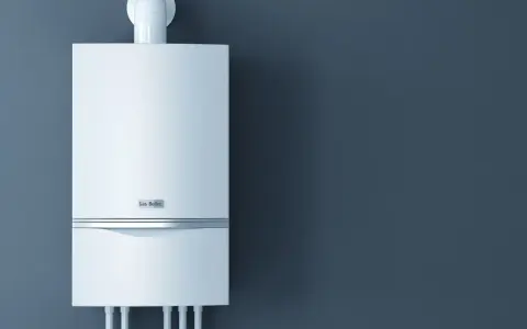 Nowoczesny piec gazowy 24kW na ile metrów zapewni ciepło? Biały, wiszący na ciemnoszarej ścianie, z widocznymi przyłączami.