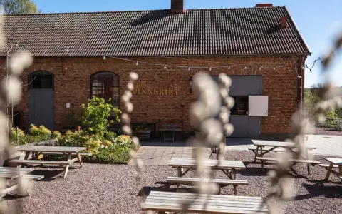 Utomhusmiljö vid Lilla Spinneriet med tegelbyggnad, trädgård och flera picknickbord.