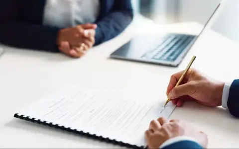 Persona firma un contrato de alquiler de habitación. A su lado, otra persona espera, con un portátil y una tablet sobre la mesa.