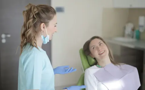 Dentystka w niebieskiej bluzie i rękawiczkach rozmawia z pacjentką. Długie studia stomatologiczne wymagają zaangażowania.