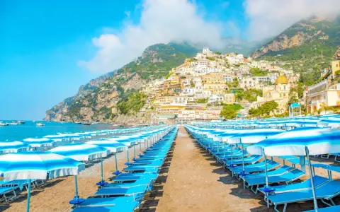 Rzędy leżaków i parasoli na plaży w Positano, Włochy. W tle widać malownicze miasteczko na zboczu góry i błękitne morze.