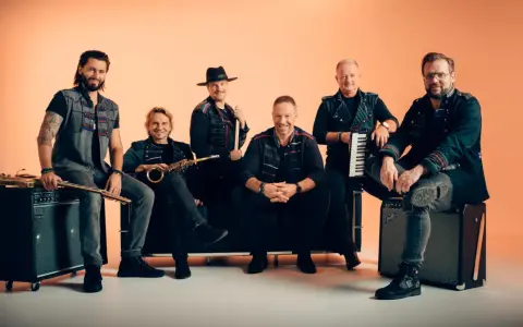 Sechs Männer einer Band posieren vor orangefarbenem Hintergrund, einige sitzen, einer steht.