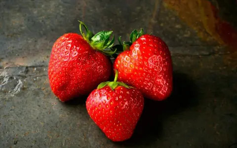 Trzy soczyste truskawki na ciemnym tle. Zastanawiasz się, ile kcal ma truskawka? To pyszna i niskokaloryczna przekąska.