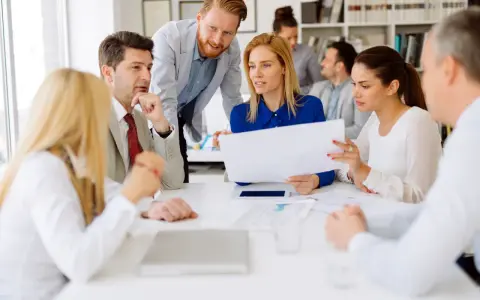 A diverse group of professionals discusses documents, possibly during board of directors recruitment or a strategic meeting.