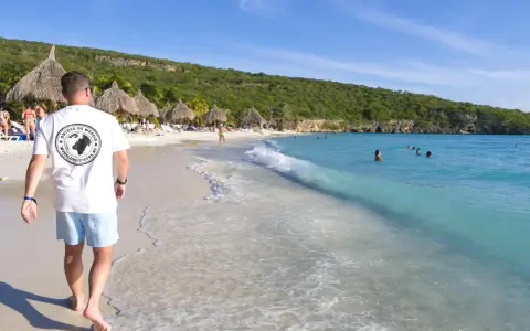En man går längs en vit sandstrand med turkost vatten. Palmklippor och parasoll syns i bakgrunden. En perfekt plats att undra var ligger Curacao.