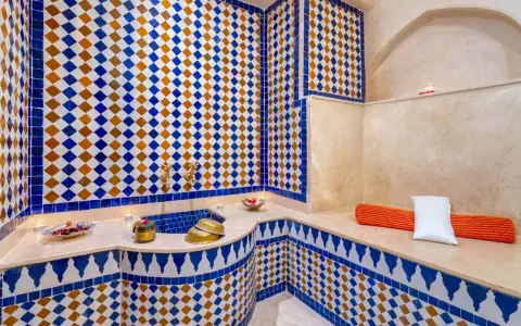 Ein luxuriöser Hamam in Marokko, verziert mit blauen und orangen Zellige-Fliesen. Kerzen und Schalen mit Rosenblättern schaffen eine entspannende Atmosphäre.