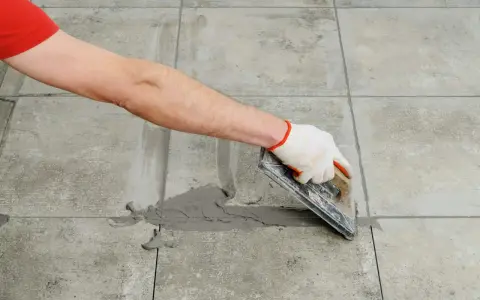 Mano con guante aplica lechada gris a las juntas entre azulejos de cemento con una llana.