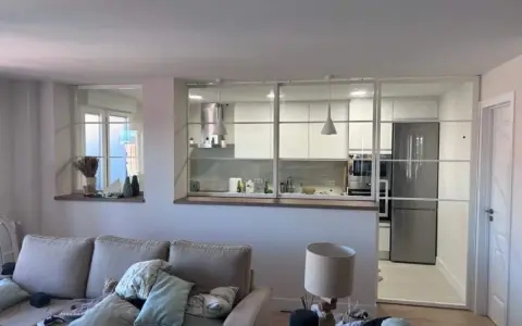 Un salón moderno con sofá y lámpara, separado de una cocina blanca y luminosa por una cristalera.