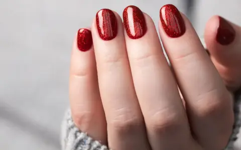 Glitzernde rote Gel-Nagel-Designs auf einer Hand, die aus einem grauen Pullover ragt.