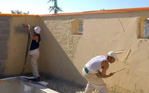 Deux ouvriers appliquent du crépi extérieur. L'un utilise un pistolet pour projeter le matériau, l'autre lisse le crépi à la truelle.