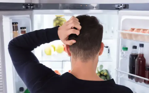Un homme se gratte la tête devant un réfrigérateur ouvert, plein de nourriture. Il semble réfléchir à un réglage frigo.