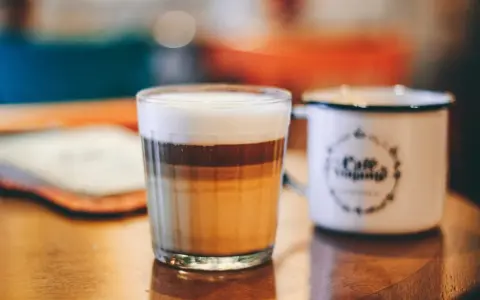 Ein geschichteter Latte Macchiato im Glas, daneben eine weiße Tasse mit Kaffee-Aufdruck.