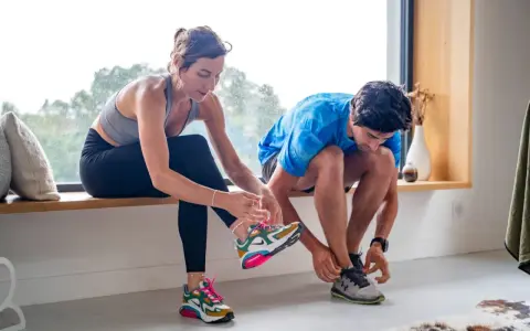 Un couple se prépare pour reprendre le sport, enfilant leurs chaussures de course près d'une fenêtre lumineuse.