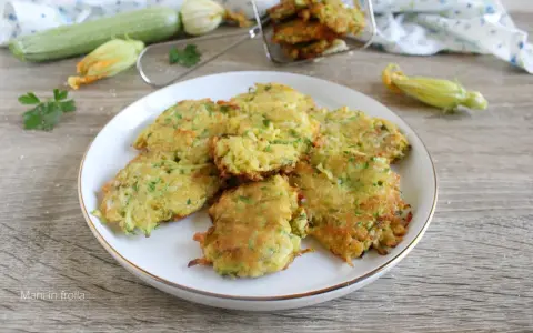Buñuelos de calabacín dorados y crujientes en un plato blanco, con calabacines y flores en el fondo.