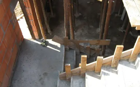 Budowa klatki schodowej: betonowe stopnie, drewniane szalunki i ceglane ściany. W tle widoczne drewniane podpory.