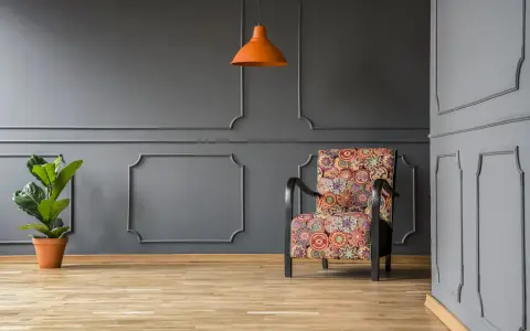 Nowoczesne sztukateria ścienna inspiracje: fotel w kolorowe wzory, pomarańczowa lampa i roślina na tle szarej ściany.