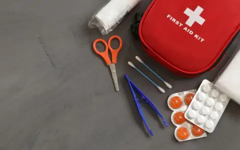 Czerwona apteczka pierwszej pomocy z krzyżem. Obok nożyczki, pęseta, waciki, bandaż i tabletki. Pamiętaj, czego nie powinna zawierać apteczka pierwszej pomocy.