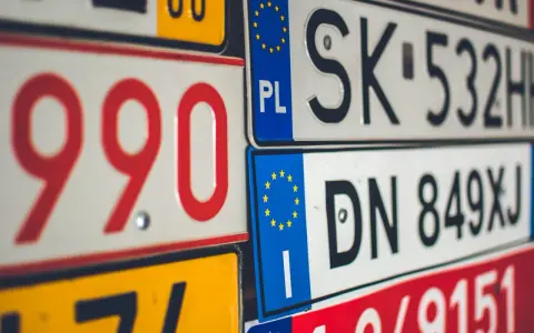 Stos tablic rejestracyjnych, w tym polskie z symbolem PL i europejską gwiazdką. Widać numery takie jak 990, SK 532HH, DN 849XJ.