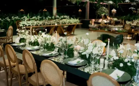 Eleganckie wesele na 100 osób. Długi, lustrzany stół z białymi kwiatami i świecami, otoczony wiklinowymi krzesłami.