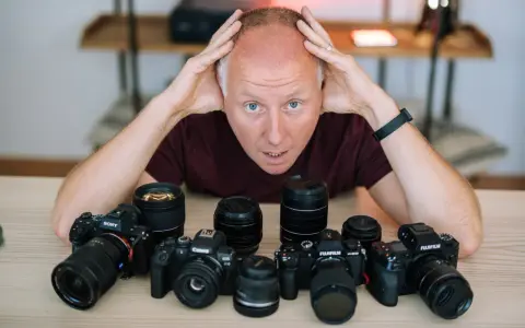 Mężczyzna z rękami na głowie otoczony aparatami różnych marek: Sony, Canon, Fujifilm.