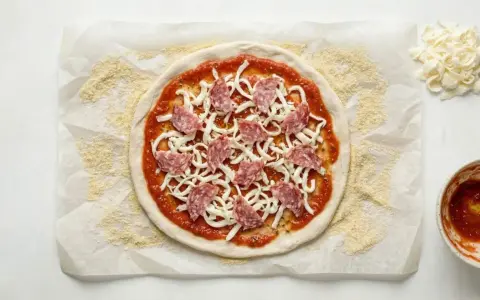 Gotowe ciasto na pizzę bez wyrastania, posmarowane sosem, posypane serem i salami. Obok miseczka sosu i dodatkowy ser.
