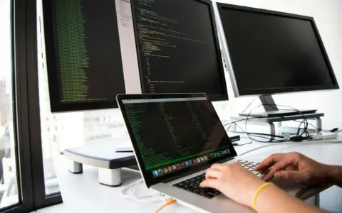 Hands typing on a laptop, surrounded by monitors displaying code. This setup is ideal for developing network infrastructure software.