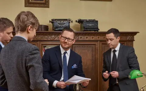 Czterech mężczyzn w garniturach rozmawia w gabinecie z zabytkowymi maszynami do pisania.