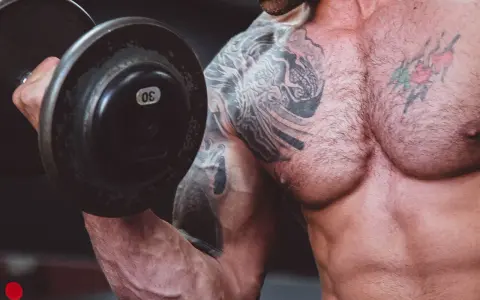 Muskulös man med tatueringar lyfter en 30 kg hantel. Frågan är om man kan bygga muskler på kaloriunderskott.