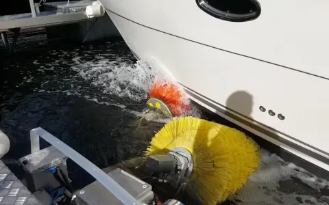 Nettoyage de la partie immergée d'un bateau avec des brosses rotatives jaunes et oranges. L'eau éclabousse autour des brosses.