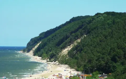 Słoneczna plaża u podnóża zalesionej Kawczej Góry. Woda faluje, a na piasku odpoczywają ludzie.