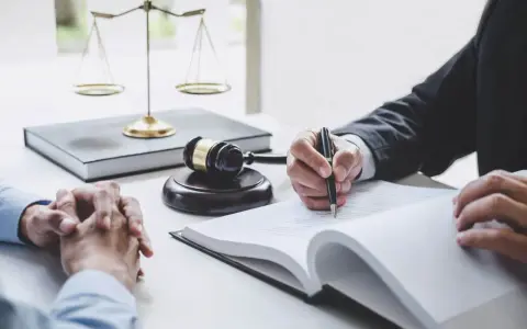 Dłoń prawnika pisząca w księdze, obok wagi sprawiedliwości i młotka sędziowskiego.