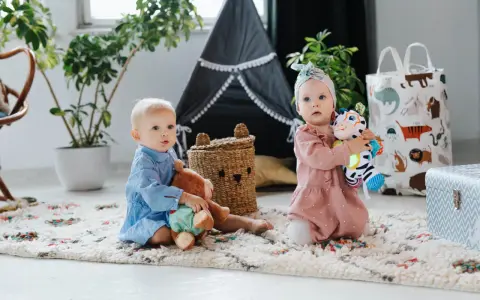 Dwoje dzieci bawi się na dywanie. Jedno trzyma misia, drugie kolorową zebrę. Idealne zabawki, co kładziemy dziecku na roczek.