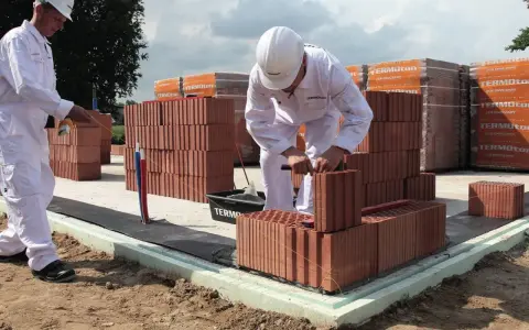 Dwóch budowlańców w białych kombinezonach układa cegły na fundamencie.