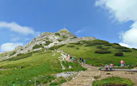 Tłum turystów wspina się kamienistym szlakiem na Giewont, podziwiając zielone zbocza i błękitne niebo.