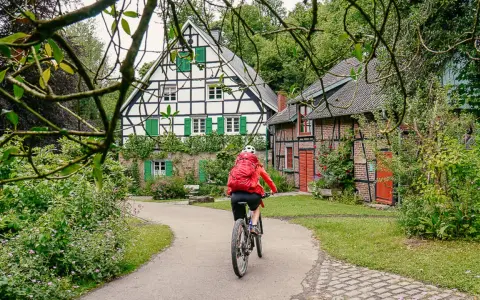 Radfahrerin auf einem Weg zwischen Fachwerkhäusern in NRW. Grüne Fensterläden und üppige Vegetation schmücken die idyllische Szenerie.