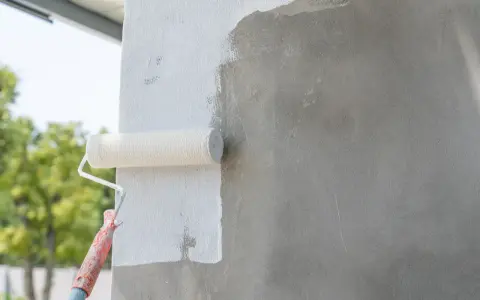 Malowanie betonu wałkiem. Biała farba pokrywa szarą ścianę, nadając jej świeży wygląd.