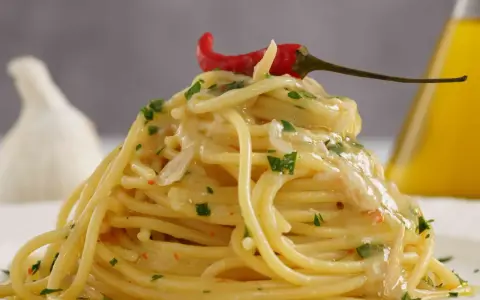 Spaghetti conditi con aglio, olio e peperoncino, guarniti con un peperoncino rosso e prezzemolo. Sullo sfondo, un bulbo d'aglio e una bottiglia d'olio.
