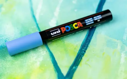 En ljusblå uni POSCA vattenfast tuschpenna ligger på en abstrakt målning i gröna och blå nyanser.