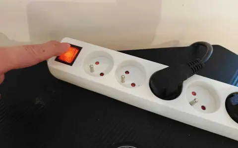 Doigt appuyant sur l'interrupteur d'une multiprise blanche, plusieurs prises vides et une prise avec un câble noir.