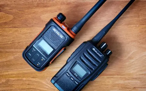 Dwa radiotelefony, jeden pomarańczowo-czarny RETCIS, drugi czarny Midland, leżą na drewnianym blacie.