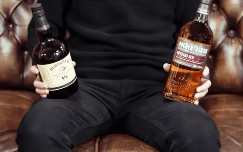 Mann hält Flaschen von Redbreast (irischer Whiskey) und Auchentoshan (schottischer Whiskey), um den Unterschied zu zeigen.