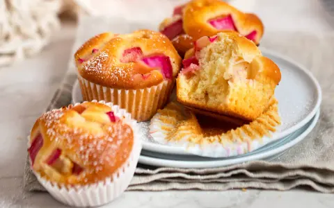 Saftige Rhabarber Muffins, mit Puderzucker bestäubt, liegen auf einem Teller. Einer ist angeschnitten und zeigt die rosa Rhabarberstücke.
