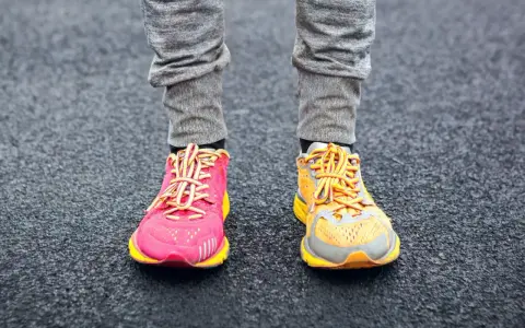 Gambe con pantaloni grigi e scarpe da corsa, una rosa acceso e l'altra arancione, con lacci gialli. La suola scarpa è pronta per correre.