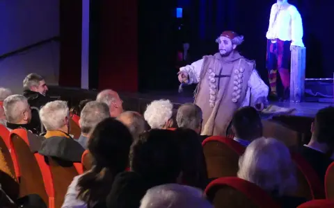 Attori sul palco, uno con un costume particolare, sembrano rompere la **quarta parete significato** parlando al pubblico.