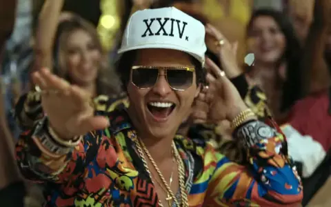 Bruno Mars w Polsce! Wesoły artysta w kolorowej koszuli i czapce z napisem "XXIV K" tańczy wśród rozbawionych ludzi.