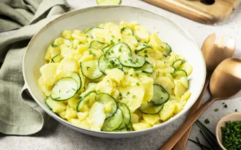 Schüssel mit Kartoffelsalat mit Gurken, garniert mit Schnittlauch. Daneben liegen Servierbesteck und eine kleine Schale mit gehacktem Schnittlauch.