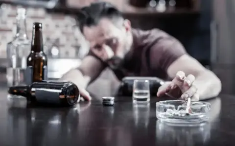 Mężczyzna z papierosem w dłoni, otoczony butelkami i popielniczką, pokazuje, czy alkohol to depresant.