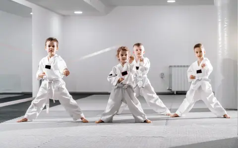 Cztery dzieci w białych kimonach ćwiczą judo dla dzieci. Stoją w pozycji bojowej na macie w jasnej sali treningowej.